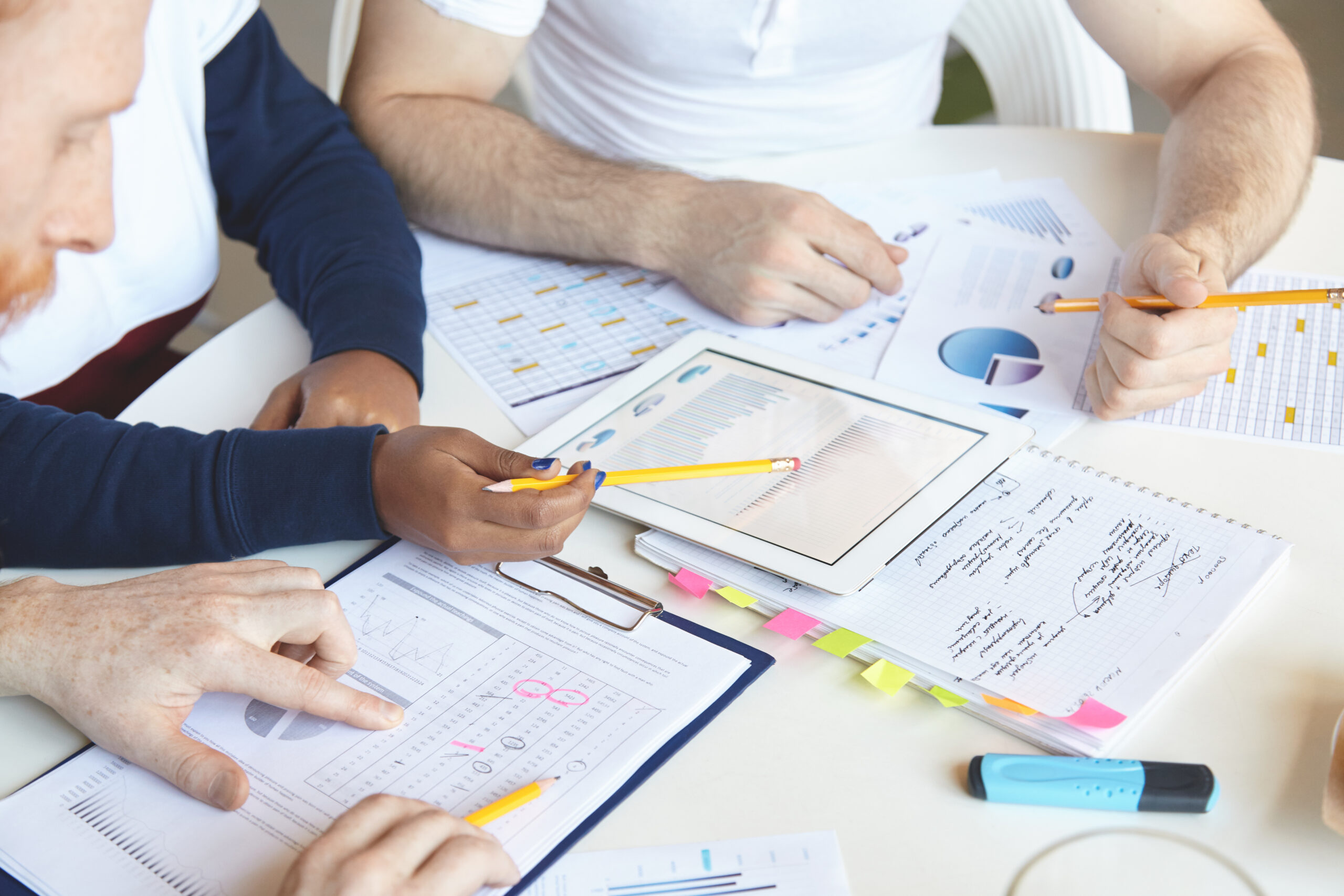 creative people of diverse ethnicities working on business plan together, analyzing growth rate, value of goods and services, studying market, counting loss, using touch pad pc and making notes